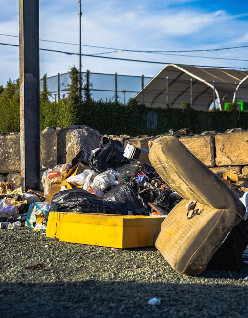 A pile of mixed waste materials and rubbish on a paved outdoor surface, including several black plastic garbage bags, a yellow plastic storage container, and an old, soiled car seat that is beige with visible stains, leaning against a stone wall made of roughly cut beige bricks. In the background, there is a tall utility pole with electrical wires running across the sky, which is partly cloudy with patches of blue. Behind the stone wall, a green hedge is visible, along with a large, semi-circular fabric-covered sports court or enclosure, supported by white metal framing. The scene appears to be part of a private waste collection or on-site rubbish clearance, consistent with the services of Waste Removal Bermondsey, situated in an industrial or semi-residential area, with natural daylight illuminating the cluttered scene, highlighting the textures of the discarded materials and surrounding environment.