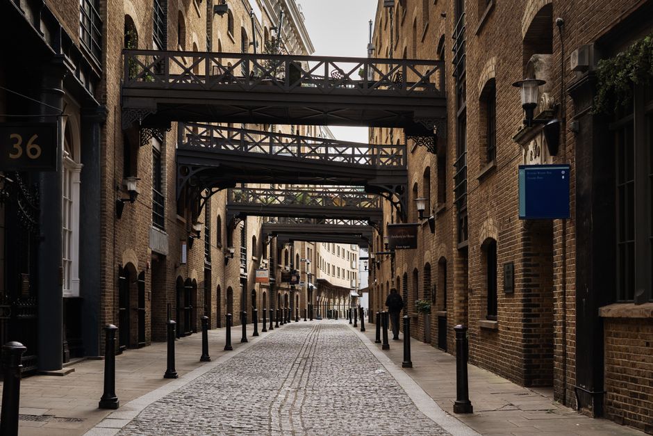 A narrow cobblestone street in an urban area is lined with multi-storey brick buildings on both sides, featuring arched windows and black metal balcony railings. Overhead, several dark, decorative metal bridges connect the buildings across the street, creating an enclosed, corridor-like atmosphere. The street is bordered by evenly spaced black bollards along the pavement, which guide pedestrian movement and provide a barrier from possible vehicle access. The scene appears quiet with minimal foot traffic, and the lighting suggests an overcast sky, resulting in soft, diffuse natural illumination that highlights the textures of the brickwork, cobblestones, and metalwork. Visible signage on the building walls indicates commercial or residential use, and no visible rubbish or waste is present in the scene, aligning with an environment where private waste management or clearance services like Waste Removal Bermondsey could operate to maintain cleanliness and accessibility in such historical urban settings.