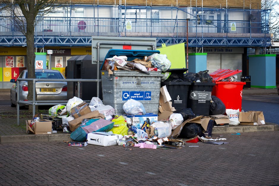 A pile of mixed waste materials and rubbish on a paved outdoor surface, including several black plastic garbage bags, a yellow plastic storage container, and an old, soiled car seat that is beige with visible stains, leaning against a stone wall made of roughly cut beige bricks. In the background, there is a tall utility pole with electrical wires running across the sky, which is partly cloudy with patches of blue. Behind the stone wall, a green hedge is visible, along with a large, semi-circular fabric-covered sports court or enclosure, supported by white metal framing. The scene appears to be part of a private waste collection or on-site rubbish clearance, consistent with the services of Waste Removal Bermondsey, situated in an industrial or semi-residential area, with natural daylight illuminating the cluttered scene, highlighting the textures of the discarded materials and surrounding environment.