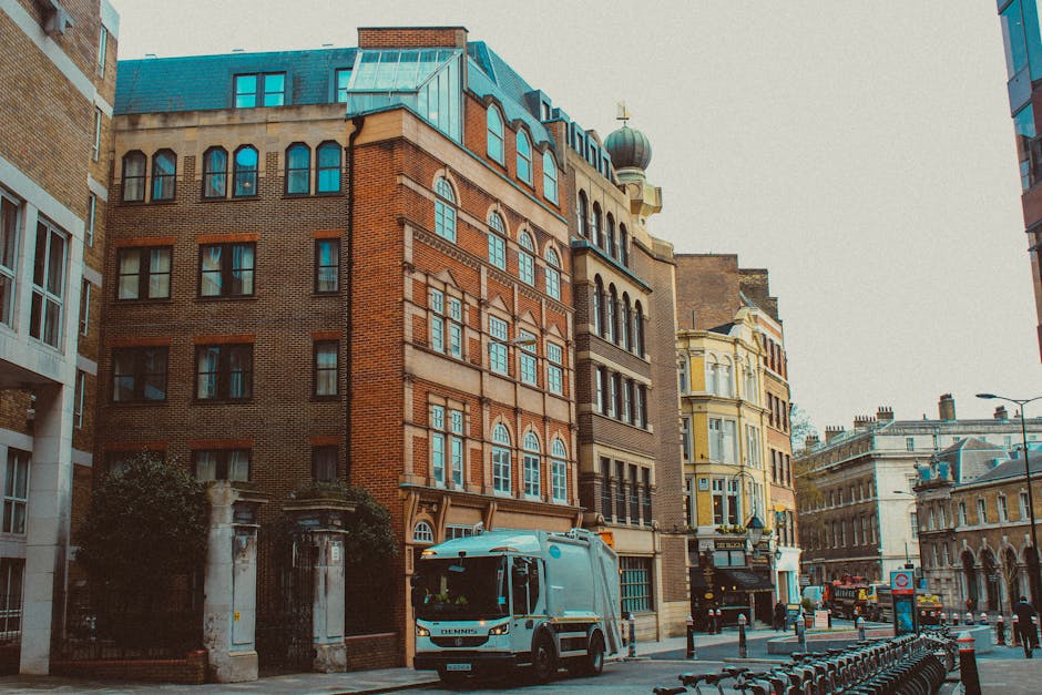 A street scene featuring a multi-storey brick building with a mix of red and brown bricks, large arched windows with white framing, and rooftop elements including a teal-colored metal roof and a domed cupola with a small golden cross. In the foreground, a light blue rubbish collection truck is parked on the pavement near a gated entrance with white stone pillars. The street is lined with bollards and a row of parked bicycles, with other historic and modern buildings visible further down the street. The environment appears to be in an urban area with a clear view of the sky, casting natural daylight across the scene. The presence of the rubbish truck indicates a private waste collection service, aligning with alternative rubbish removal methods.