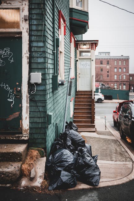 A narrow urban alleyway features a building with weathered teal wooden cladding and red-trimmed windows. At the base of the building, several black trash bags are piled on the concrete ground, near a small strip of dried grass and a concrete staircase with wooden steps leading up to a white door. A white utility box and electrical cables are mounted on the building’s exterior, with some graffiti visible on an adjacent door frame. The background shows a parking lot with cars and a row of brick buildings under an overcast sky. The scene illustrates a typical example of waste awaiting collection in a city environment, with Waste Removal Bermondsey perhaps providing private rubbish removal services for residents seeking alternative waste handling options outside local authority collection. The overall composition emphasizes the practical aspects of rubbish storage outside residential properties, with a focus on the clutter of refuse bags and the surrounding urban setting, captured during daylight with diffused natural light evident in the muted tones and textures of the scene.
