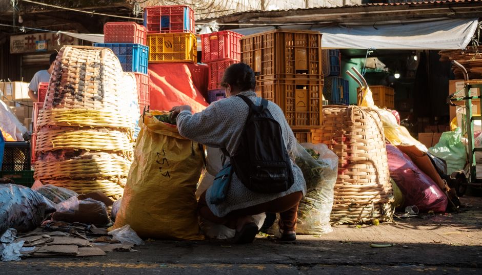 A person kneeling on the ground at an outdoor market or alleyway during daylight, surrounded by large stacks of woven baskets, plastic crates, and bags filled with various types of waste or discarded materials. The baskets are made of natural, light-colored woven fibers, with some stacked neatly and others leaning or partially collapsed. The plastic crates are in shades of red, blue, yellow, and brown, and are arranged in piles behind the person, with some crates showing signs of wear or dirt. Large yellow and pink plastic bags, possibly filled with rubbish or recyclables, are situated near the individual, who is wearing a grey sweater, dark pants, and has a black backpack. The ground is paved and scattered with smaller debris, including crumpled paper and plastic waste. In the background, there are some structures with corrugated metal roofing and tarpaulins, indicating a marketplace or informal waste collection area. The scene appears to be in natural light, with some shadowed areas, highlighting the textures and colors of the waste materials, and reflecting the context of independent waste handling or rubbish removal services in a busy outdoor setting.