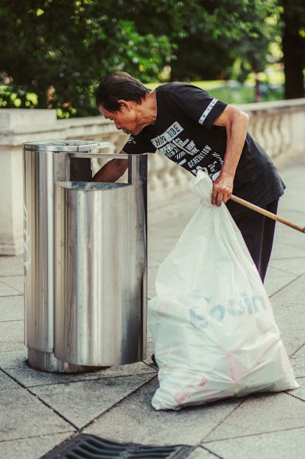 A middle-aged woman with short dark hair is seen disposing of rubbish into a stainless steel public trash bin on a paved walkway. She is wearing a black T-shirt with white printed text on the front, and is bent slightly forward as she places a white plastic bag filled with waste into the bin. Her left hand holds the bag steady, while her right hand uses a wooden stick to assist in guiding the rubbish into the container. The trash bin is cylindrical with a shiny, reflective surface and an open top, positioned in front of a low stone railing. In the background, lush green trees and a well-maintained park or garden area are visible, suggesting an outdoor urban environment. The scene is illuminated by natural daylight, indicating a clear day. This image demonstrates a community-based waste disposal activity, aligning with private rubbish removal or on-site collection services such as those provided by Waste Removal Bermondsey, in a typical outdoor setting suited to rubbish management activities.
