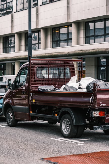 A small burgundy flatbed truck parked on a city street, filled with a variety of waste materials including black plastic bags, cardboard boxes, and loose debris. The truck features a metal mesh safety partition behind the cab, with some of the waste resting on the truck bed's wooden floor and side panels. The vehicle's rear lights and wheel arches are visible, and the truck is positioned next to a modern office building with large windows and a light-colored facade. In the background, other parked vehicles and urban infrastructure, such as a streetlamp, can be seen. The scene appears to be during daylight hours with natural light illuminating the surroundings, indicative of an ongoing or completed rubbish removal operation facilitated by an independent waste management service like Waste Removal Bermondsey, suitable for private collection or on-site clearance needs in an urban environment.