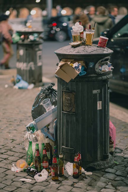 A black public rubbish bin made of metal with a textured, ridged surface is situated on a cobbled sidewalk. The bin is overflowing with various discarded items, including crumpled food packaging, paper boxes, and plastic bags, some of which hang loosely from the sides. Several glass bottles, primarily beer bottles with green and brown tint, are scattered at the base, some partially crushed or with caps removed. On top of the bin, there are leftover food wrappers, a plastic cup with a lid and straw, and a disposable coffee cup, indicating nearby casual outdoor dining or social activity. In the background, a blurred scene features parked cars, pedestrians, and an additional waste container, suggesting an urban street environment. The atmosphere appears to be daytime, with natural light illuminating the scene, and the area reflects typical urban littering associated with inconsistent rubbish disposal practices, despite the availability of public waste collection points. Waste Removal Bermondsey and other private rubbish services often handle such overflow or street cleaning tasks in these areas, addressing local waste management needs in accordance with municipal regulations.
