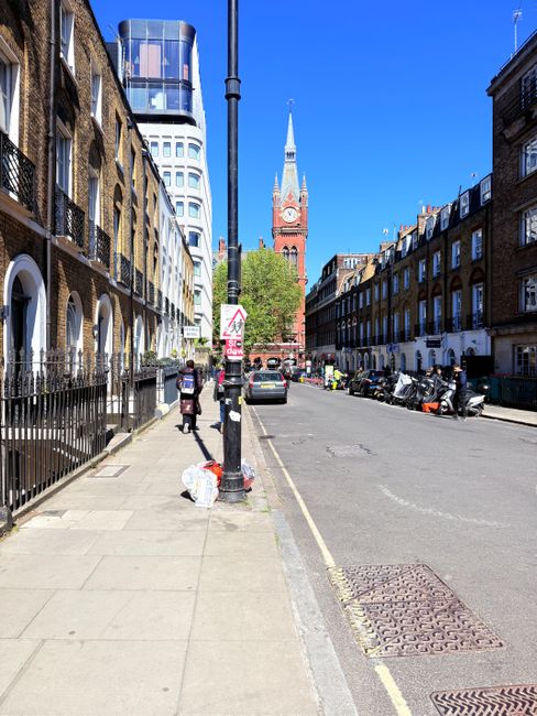 A wide urban street scene in Bermondsey, with a clear blue sky overhead. The foreground features a paved sidewalk with a few pedestrians, some of whom are carrying bags, walking past black metal railings in front of row houses with brick facades and white-framed windows. A black pole with a warning sign and a small pile of white rubbish bags are positioned on the sidewalk near the curb. Parked motorcycles and a small vehicle are lined along the street on the right side, adjacent to a yellow double line marking the edge of the parking zone. In the background, a prominent red brick clock tower with a tall, pointed spire and a clock face dominates the skyline, flanked by modern glass-fronted buildings. The overall scene reflects a typical city street with mixed residential and commercial architecture, where private waste disposal in rubbish bags is likely part of local rubbish management practices, supported by services such as Waste Removal Bermondsey.